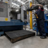 In a brightly lit factory, a worker stands on an Interlocking Anti-Fatigue Mat while operating an industrial machine, holding a tool near its open compartment. The area is organised with visible equipment and safety gear.