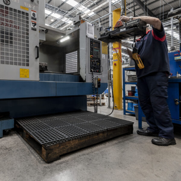 In a brightly lit factory, a worker stands on an Interlocking Anti-Fatigue Mat while operating an industrial machine, holding a tool near its open compartment. The area is organised with visible equipment and safety gear.