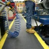A person in jeans and tan work boots stands on a factory floor, positioned next to machinery and coiled blue air hose, standing on a Deckplate Anti-Fatigue Mat bordered by yellow safety lines marking the walkway.
