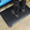 A person in black trousers and trainers stands on an Anti-Static Deckplate Anti-Fatigue Mat with a yellow lead, placed on concrete by a blue metal shelf.