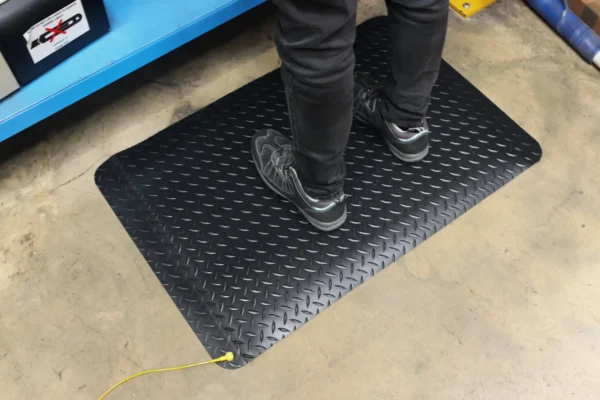 A person in black trousers and trainers stands on an Anti-Static Deckplate Anti-Fatigue Mat with a yellow lead, placed on concrete by a blue metal shelf.