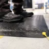 Close-up of a person standing on an Anti-Static Deckplate Anti-Fatigue Mat with a yellow earthing cord on a concrete floor in an industrial setting. Only lower legs and feet in black shoes are visible.