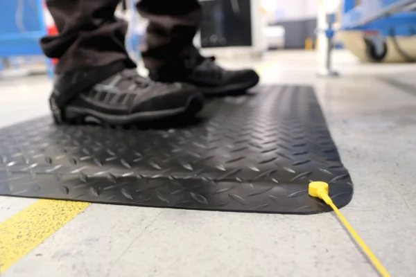 Close-up of a person standing on an Anti-Static Deckplate Anti-Fatigue Mat with a yellow earthing cord on a concrete floor in an industrial setting. Only lower legs and feet in black shoes are visible.