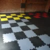 Black Circular Stud Interlocking Tiles are arranged in chequerboard patterns with yellow, red, and grey sections. A brick wall appears in the background.