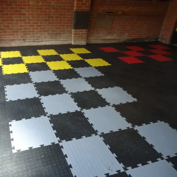Black Circular Stud Interlocking Tiles are arranged in chequerboard patterns with yellow, red, and grey sections. A brick wall appears in the background.