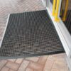 A Cross-Ribbed Entrance Mat in black is placed outside a glass door with yellow handles, resting on a brick-patterned walkway.