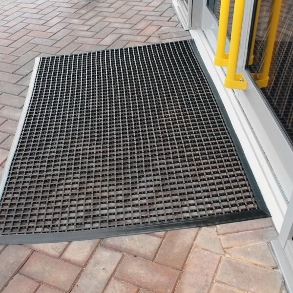A Cross-Ribbed Entrance Mat in black is placed outside a glass door with yellow handles, resting on a brick-patterned walkway.