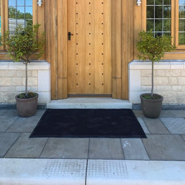 A light wooden door with decorative metal studs, flanked by two small potted trees, sits at the entrance of a stone building. A large Cross-Ribbed Entrance Mat lies in front of the step on a tiled patio.