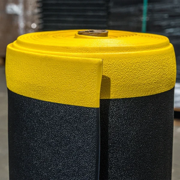 A close-up of the ESD Textured Anti-Fatigue Mat, with a yellow top and black bottom, standing upright in a warehouse.