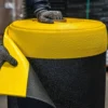Wearing gloves, a person lifts the edge of an ESD Textured Anti-Fatigue Mat, showing its yellow top layer and black textured bottom layer.