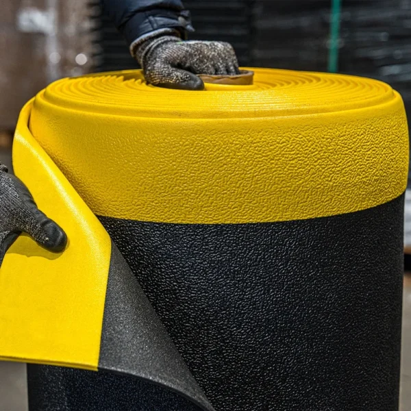 Wearing gloves, a person lifts the edge of an ESD Textured Anti-Fatigue Mat, showing its yellow top layer and black textured bottom layer.
