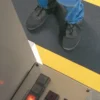 A person in black shoes and blue jeans stands on an ESD Textured Anti-Fatigue Mat with a yellow border near a metal control panel with buttons.