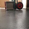 Close-up of a sack truck with red wheels on a Heavy-Duty Interlocking Tile floor in a warehouse, with cardboard boxes and a metal object visible in the background.
