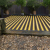 Anti-Slip Decking Strips in yellow and black are laid between wooden planks, bordered by potted trees along a dark fence, with pebbles in the foreground.