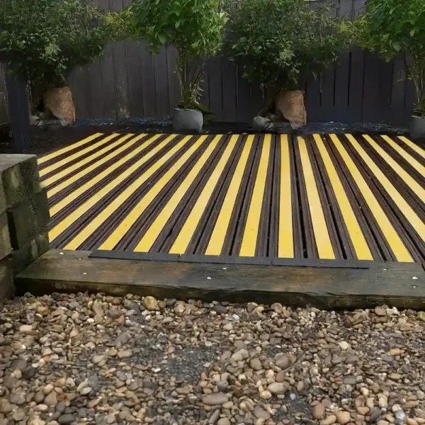A garden deck features Anti-Slip Decking Strips in alternating yellow and dark brown, bordered by potted trees along a dark fence, with a pebble path in front.