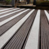 Close-up of a wooden deck featuring Anti-Slip Decking Strips in textured white, alternating with dark brown timber planks to form a striped pattern. The background is softly blurred, with hints of outdoor objects and greenery visible.
