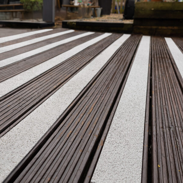 Close-up of a wooden deck featuring Anti-Slip Decking Strips in textured white, alternating with dark brown timber planks to form a striped pattern. The background is softly blurred, with hints of outdoor objects and greenery visible.