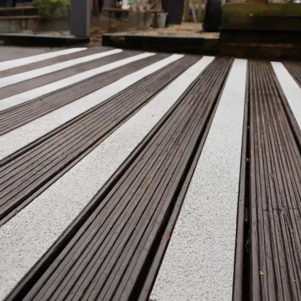 Close-up of dark wooden decking fitted with Anti-Slip Decking Strips in textured white, running parallel across the outdoor surface. The background features a blurred garden scene.