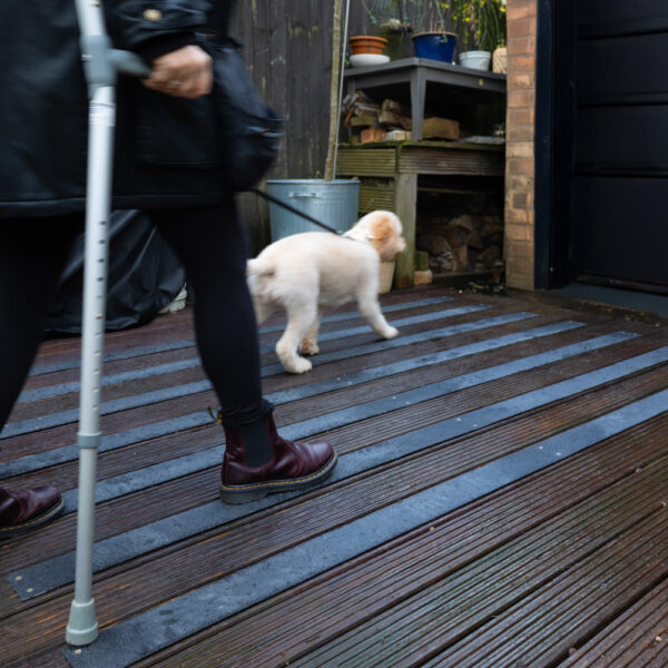 A person with a crutch and dark boots walks on a wooden deck fitted with Anti-Slip Decking Strips, following a fluffy white dog towards a doorway. Gardening items and stacked wood are visible in the background.