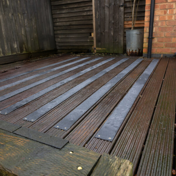 Wet and weathered wooden patio decking features several Anti-Slip Decking Strips attached in parallel lines, positioned beside a brick wall, a wooden fence, and a metal bin.
