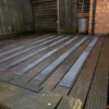 Wooden decking featuring alternating Anti-Slip Decking Strips, bordered by a wooden fence and brick wall. A metal bucket with a plant sits in the corner where the walls meet.