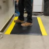 A person in brown work boots and black trousers stands on a Premium Bubble Top Anti-Fatigue Mat, which features a black surface with yellow edges, in a workshop setting.