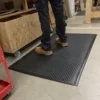A person in black trousers and tan work boots stands on a Premium Bubble Top Anti-Fatigue Mat in a workshop with wooden furniture and tools nearby.