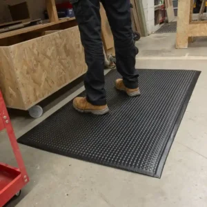A person in black trousers and tan work boots stands on a Premium Bubble Top Anti-Fatigue Mat in a workshop with wooden furniture and tools nearby.