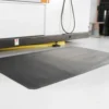A Fine Ribbed Anti-Fatigue Mat in black is positioned in front of industrial equipment on a white floor within a workspace or factory.