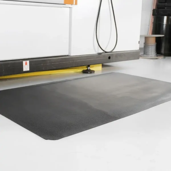 A Fine Ribbed Anti-Fatigue Mat in black is positioned in front of industrial equipment on a white floor within a workspace or factory.