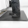 A person in black trousers and shoes stands on a Fine Ribbed Anti-Fatigue Mat, which has a textured black surface and is placed on a light-coloured floor.