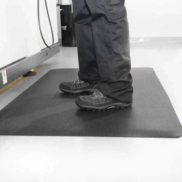 A person in black trousers and shoes stands on a Fine Ribbed Anti-Fatigue Mat, which has a textured black surface and is placed on a light-coloured floor.