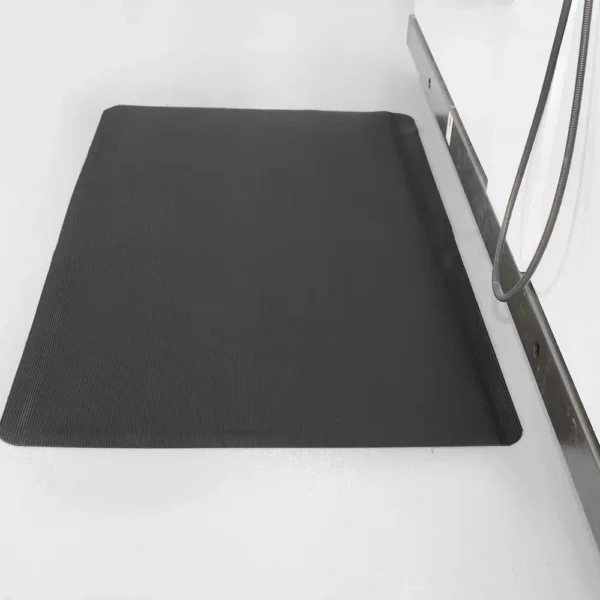 A Fine Ribbed Anti-Fatigue Mat in black is set on a light floor beside a metal workbench with a coiled cable hanging from the side.