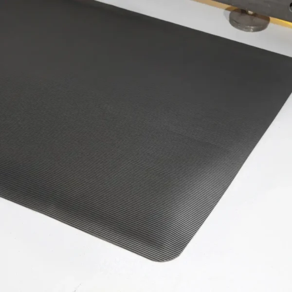 A close-up view of the Fine Ribbed Anti-Fatigue Mat in black, featuring slightly rounded corners on a white floor, with part of a metallic object visible at the top right edge.
