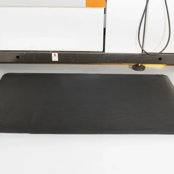 A Fine Ribbed Anti-Fatigue Mat in rectangular black is placed on a light-coloured floor before industrial equipment, with cables and a metal support bar visible in the background.