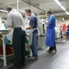 Several people wearing aprons work at assembly stations in a well-lit factory, standing on Textured Anti-Fatigue Mats as they handle tools and materials. In the background, one person in casual clothes and shorts is also present.