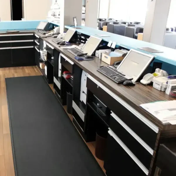 A row of modern desks with computers and office supplies sits in a bright room with wooden flooring, blue accents, and empty chairs; Textured Anti-Fatigue Mats are placed underfoot for comfort.