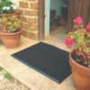 A Flexible Bristle Mat in black is at the entrance, surrounded by terracotta pots with pink and red flowering plants. The door is open, showing shoes on a tiled floor inside.