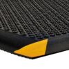 A close-up of the Foam Cushioned Industrial Mat shows its black, textured surface with raised circular bumps and a peeled corner revealing a yellow foam layer underneath.