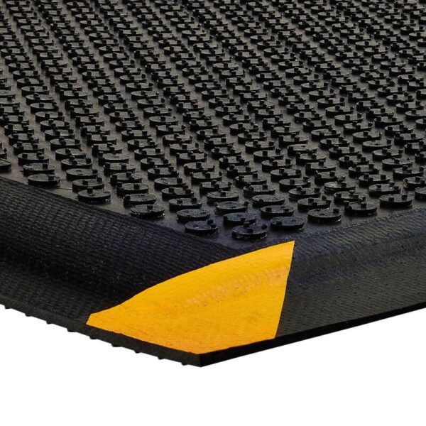A close-up of the Foam Cushioned Industrial Mat shows its black, textured surface with raised circular bumps and a peeled corner revealing a yellow foam layer underneath.