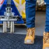 A person in blue trousers and tan work boots stands next to the wheel of a blue pallet truck on a Forklift Mat in an industrial setting.