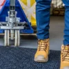 A person in blue trousers and tan work boots stands on a Forklift Mat beside a blue pallet truck and a large yellow crate in an indoor industrial setting.