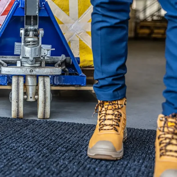 A person in blue trousers and tan work boots stands on a Forklift Mat beside a blue pallet truck and a large yellow crate in an indoor industrial setting.