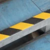A GRP Grit Tape metal step, featuring black and yellow diagonal warning stripes, is installed on a blue floor to visibly highlight and reduce trip hazards.