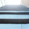 Close-up of blue tiled stairs fitted with GRP Grit Tape on each step, ascending towards a sunlit area with a glass railing on the left and a brick wall on the right.