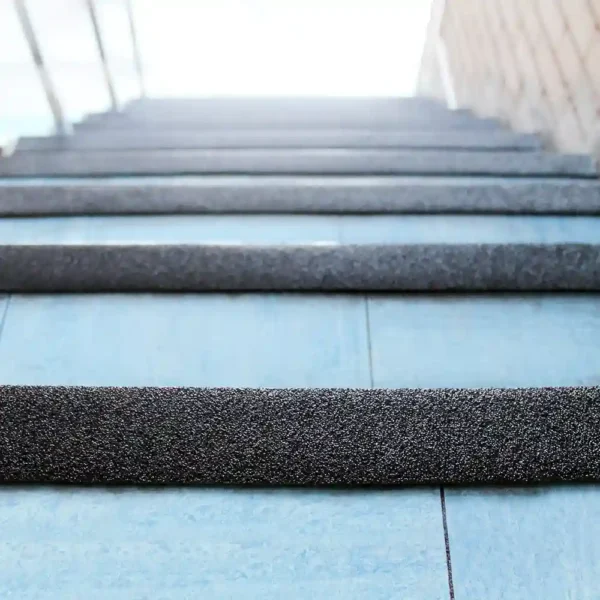 Close-up of blue tiled stairs fitted with GRP Grit Tape on each step, ascending towards a sunlit area with a glass railing on the left and a brick wall on the right.