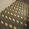 A set of carpeted stairs features yellow and black GRP Grit Tape with diagonal stripes on each step, placed against a white painted wall.