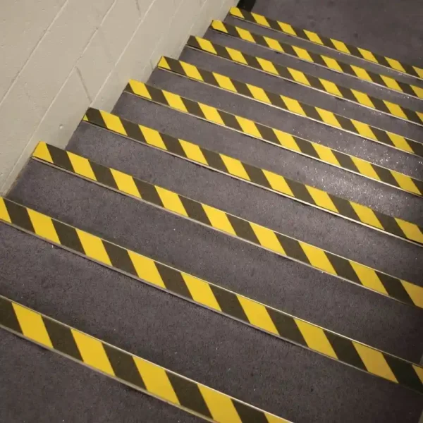 A set of carpeted stairs features yellow and black GRP Grit Tape with diagonal stripes on each step, placed against a white painted wall.