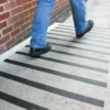 A person wearing blue jeans and black shoes walks on a concrete pavement with evenly spaced black GRP Grit Tape strips, next to a red brick wall.