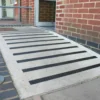 A concrete wheelchair-accessible ramp with GRP Grit Tape anti-slip strips leads to a building entrance with an open door, bordered by a red brick wall and featuring a metal handrail on one side.